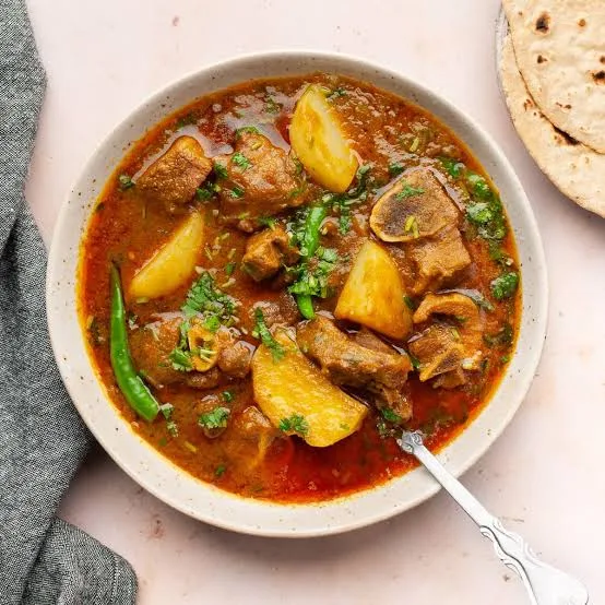 A pot of Pakistani aloo gosht with mutton pieces and potatoes in a rich red-brown gravy, garnished with coriander and ginger, served with roti.