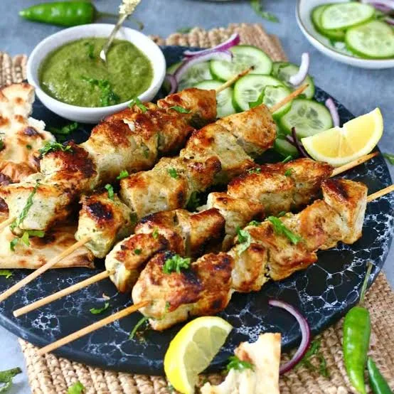 Skewers of creamy grilled chicken malai boti with lemon wedges, onion rings and green chutney on a slate board