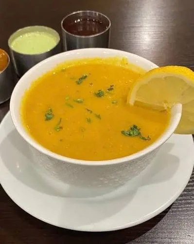 Bowl of smooth golden lentil soup topped with cumin and garlic tarka, coriander and a lemon wedge