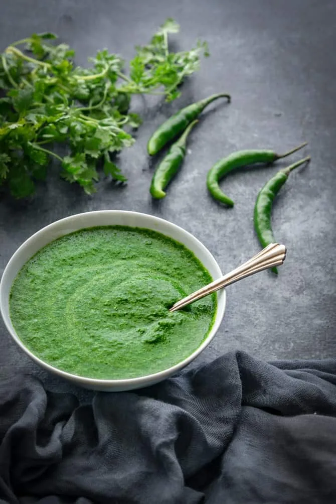 Bowl of bright green hari chutney with fresh coriander, green chillies and lemon on a wooden board