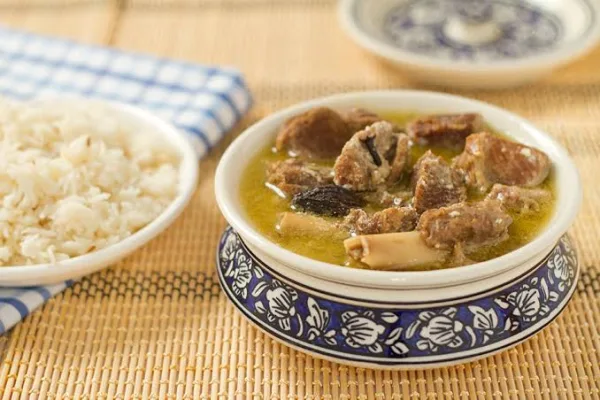 A pot of creamy pale Kashmiri yakhni with mutton pieces, whole spices and a bowl of white rice on a wooden table