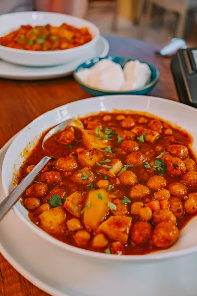 A brass bowl of dark Lahori chickpea curry topped with ginger and coriander, served with puri and lemon wedges