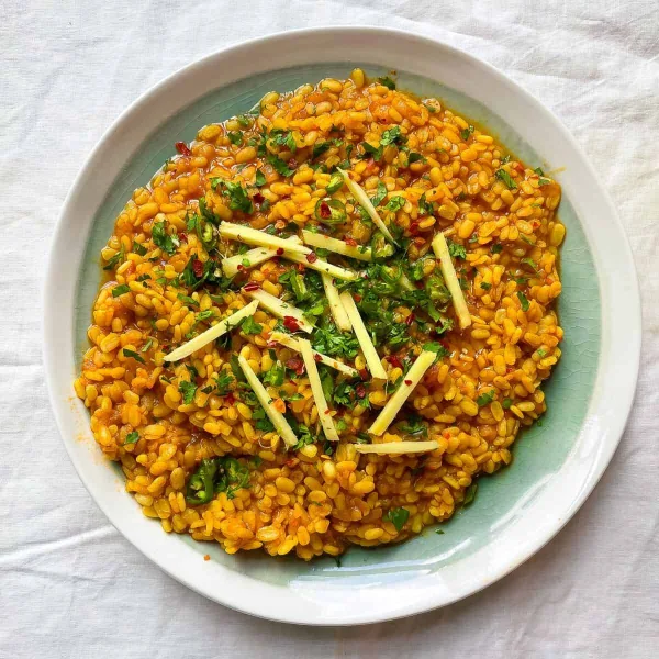 A brass bowl of creamy mash ki daal topped with garlic ghee tarka, red chillies and fresh coriander, served with roti and lemon wedges
