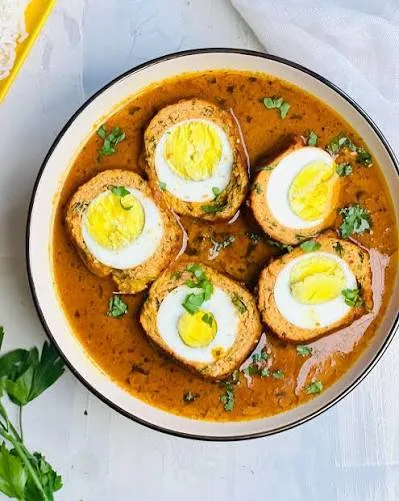Halved nargisi koftas showing boiled egg centres in a glossy orange-brown gravy, garnished with green chillies and ginger