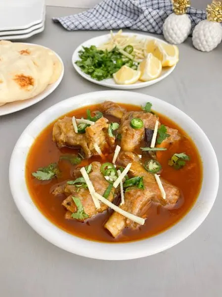 Bowl of Pakistani paya with tender trotters in a glossy spiced gravy, garnished with ginger, green chillies and coriander, served with naan and lemon.