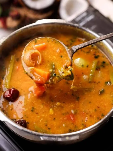 A bowl of sambar with vegetables and curry leaves, served with rice and idli
