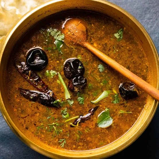A bowl of red-orange tomato tamarind rasam topped with curry leaves and coriander, served with rice and a papad